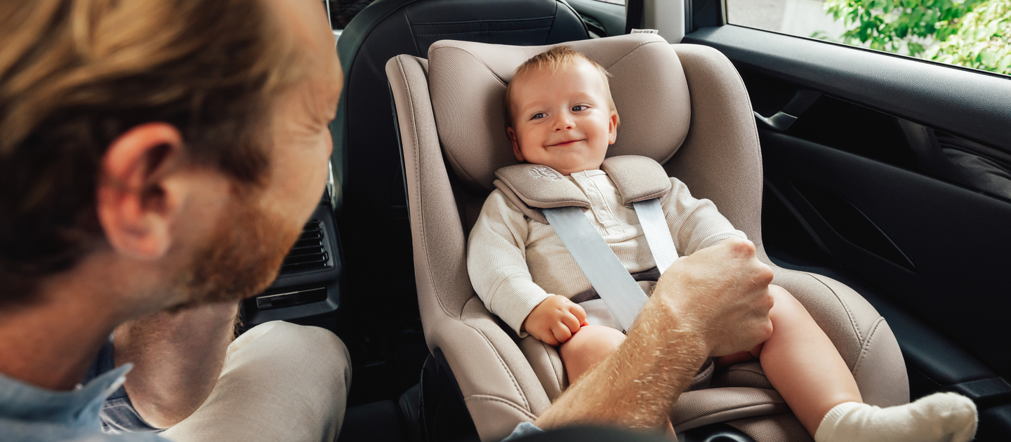 Ein l&auml;chelndes Baby sitzt sicher in einem Britax R&ouml;mer Dualfix 5Z Autositz in der Farbe Teak, der auf dem R&uuml;cksitz eines Autos montiert ist. Ein Erwachsener, von der Seite zu sehen, h&auml;lt sanft das Bein des Babys und interagiert mit ihm.